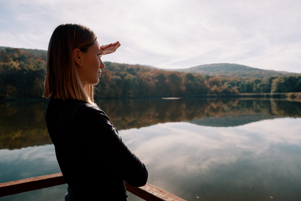 Uma pessoa está em um deck à beira de um lago calmo, protegendo os olhos enquanto olha para as colinas distantes sob um céu parcialmente nublado, refletindo sobre sua jornada e sua nova auto-estima.