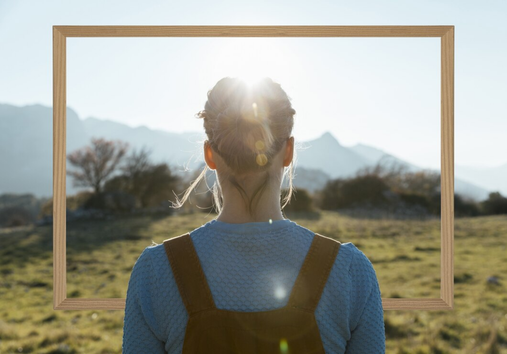 Uma pessoa com o cabelo preso para trás está em um campo, olhando com confiança para as montanhas emolduradas por uma moldura de madeira. O sol brilha intensamente sobre o topo das montanhas, iluminando a cena como um retrato de autoestima.
