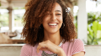 Uma mulher com longos cabelos pretos e cacheados está sorrindo ao ar livre, com as mãos juntas à sua frente. Ela está vestindo uma camisa e tem uma expressão feliz no rosto.