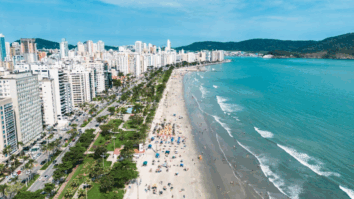 Vista aérea da praia de Santos em São Paulo, com mar azul, areia movimentada por banhistas e guarda-sóis, jardins litorâneos arborizados e prédios altos ao fundo em um dia ensolarado