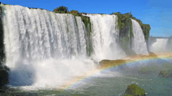 Vista panorâmica das Cataratas do Iguaçu, com quedas d’água imponentes cercadas por vegetação e um arco-íris formado pela névoa sobre o rio